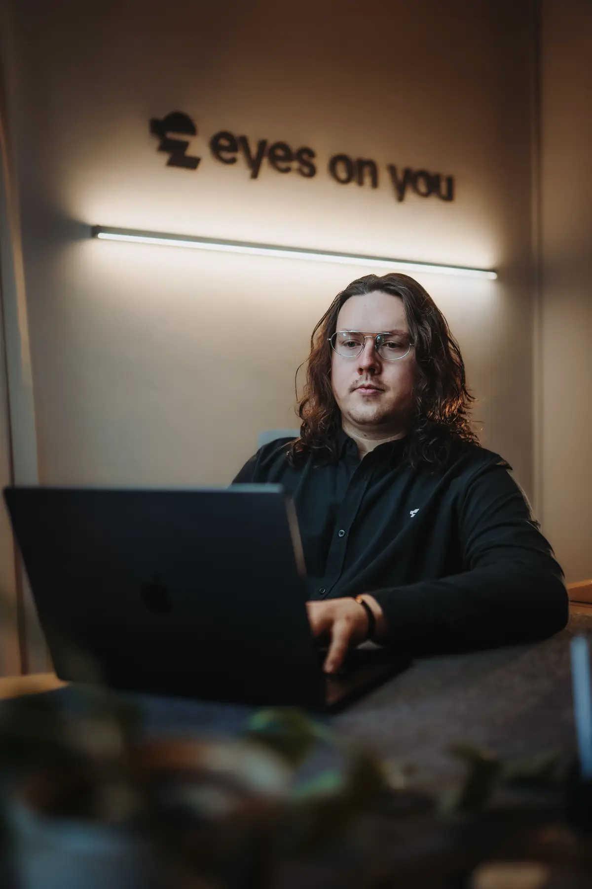 Kenan Adam arbeitet am Laptop unter dem beleuchteten Logo von eyes on you