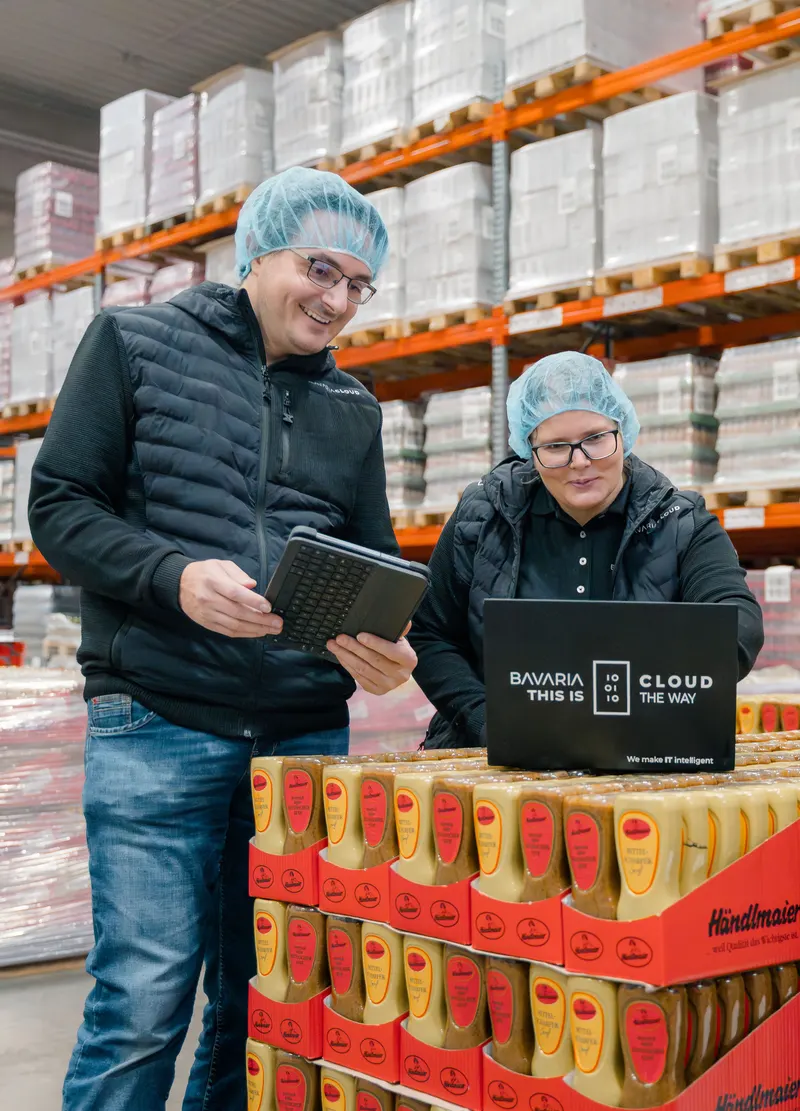 Zwei Mitarbeiter von BavariaCloud mit Haarnetz und Tablet beim Kunden Händlmaier im Lager, vor ihnen ein Laptop mit „Bavaria this is Cloud the way“