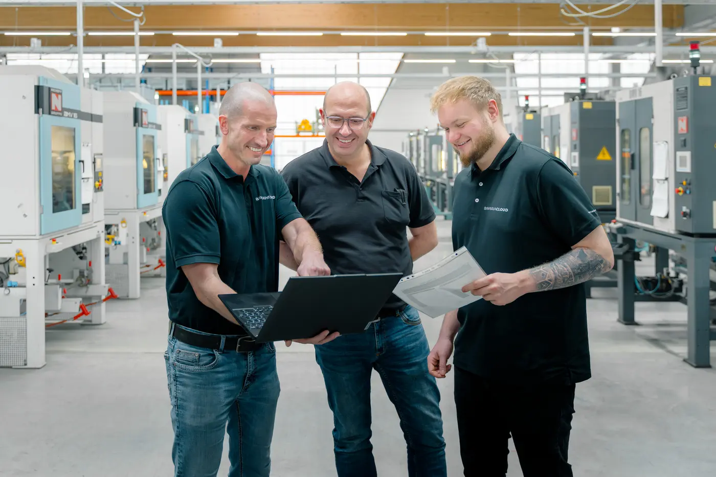 Drei Mitarbeiter von BavariaCloud lachen gemeinsam über einen Laptop gebeugt in einer Produktionshalle
