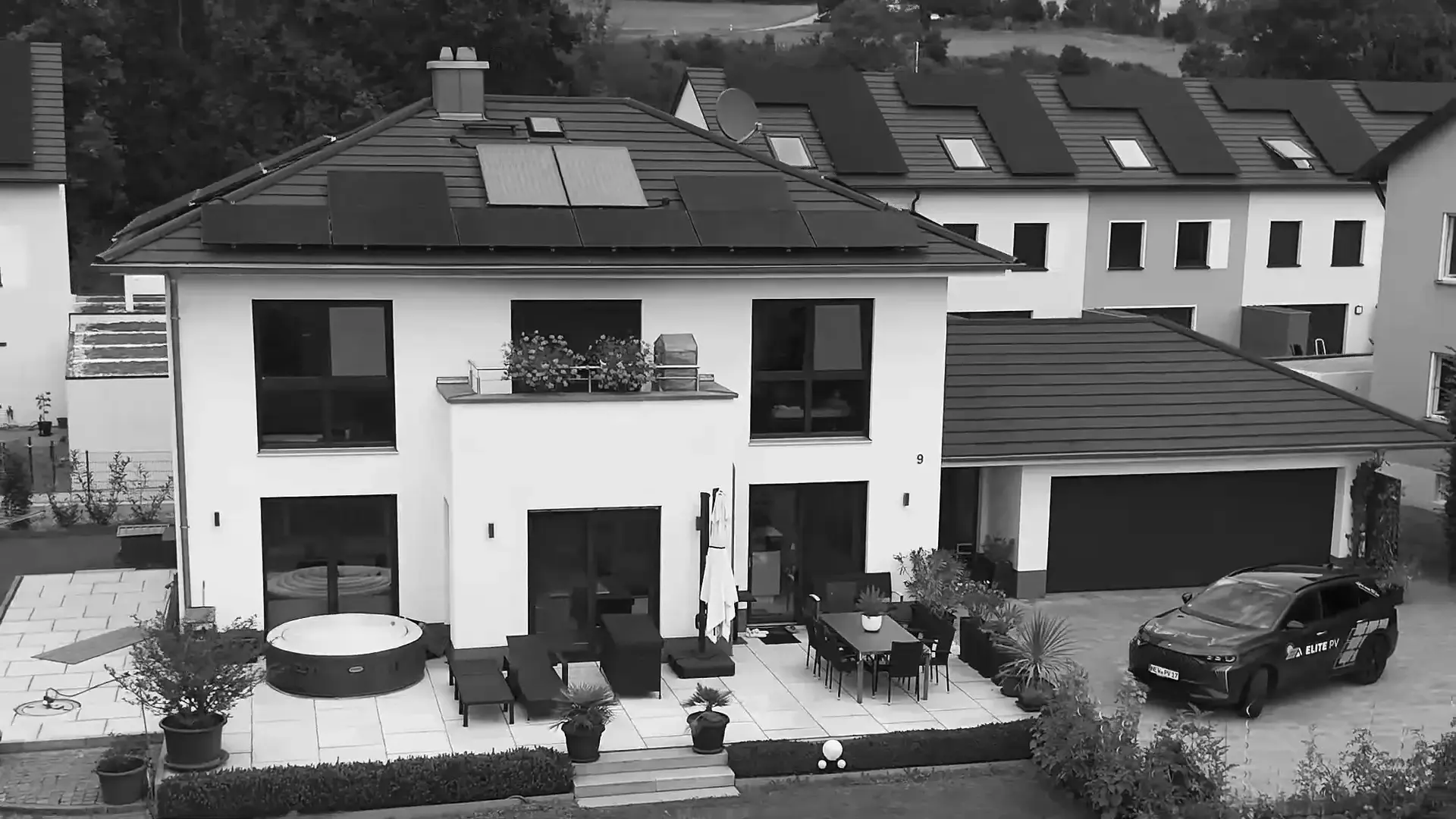 Einfamilienhaus mit Photovoltaik auf dem Dach und dem Firmenwagen von Elite PV in der Einfahrt, aus der Luft, schwarzweiß