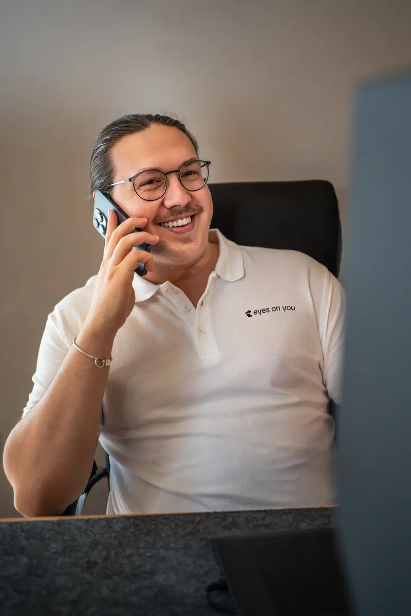 Kenan Adam lacht am Telefon an seinem Schreibtisch, im weißen Poloshirt mit eyes-on-you-Logo