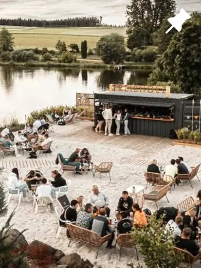 Instagram-Beitrag: Sandstrand mit Lounge-Möbeln und Gästen vor der Bar am See