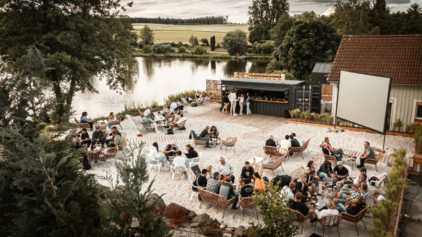 Luftaufnahme der Treibholz Beachbar am Naturbadesee Erlenweiher: Sandstrand, Liegestühle und viele Gäste am Abend