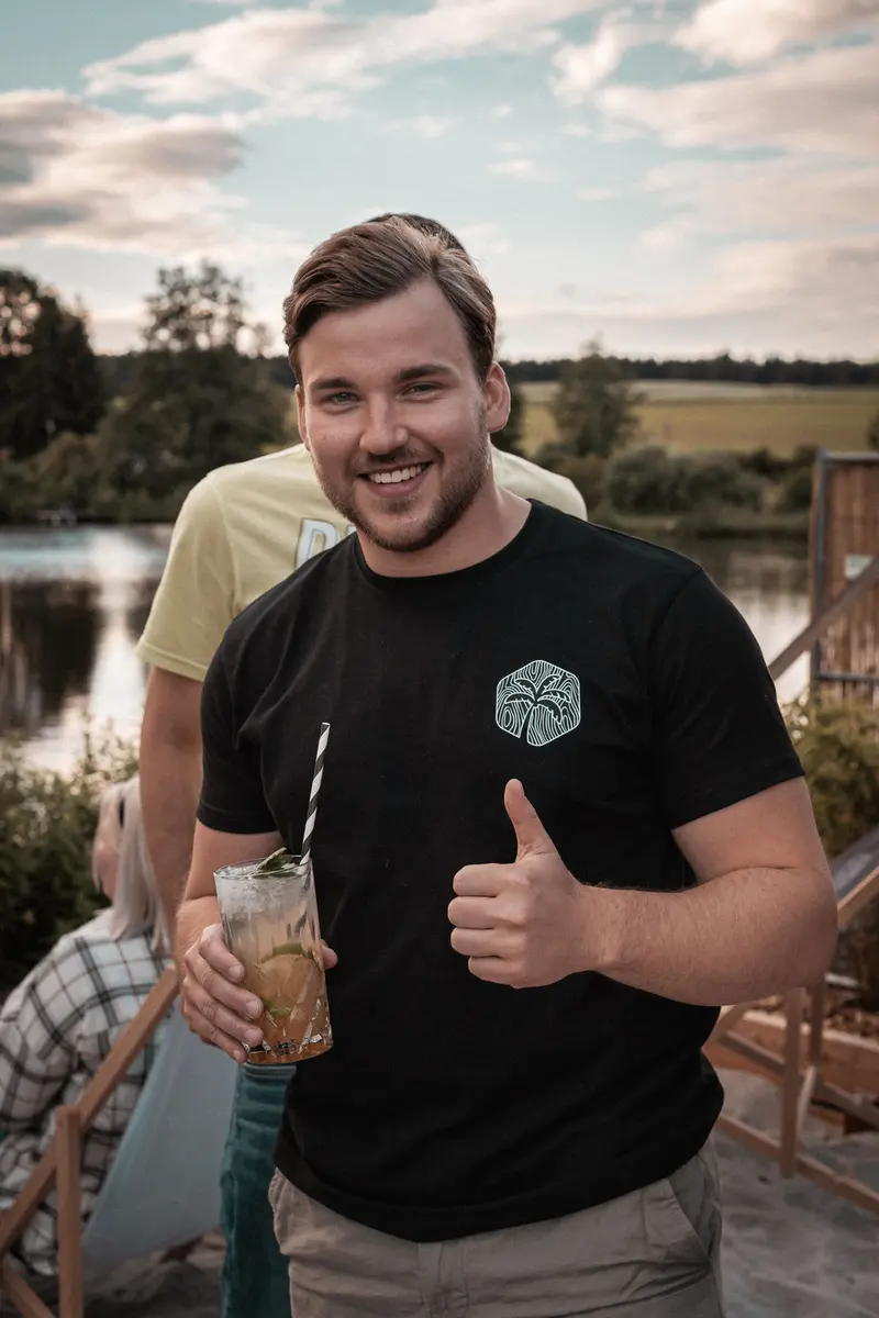 Mitarbeiter der Treibholz Beachbar mit Drink zeigt den Daumen nach oben, im Hintergrund der See